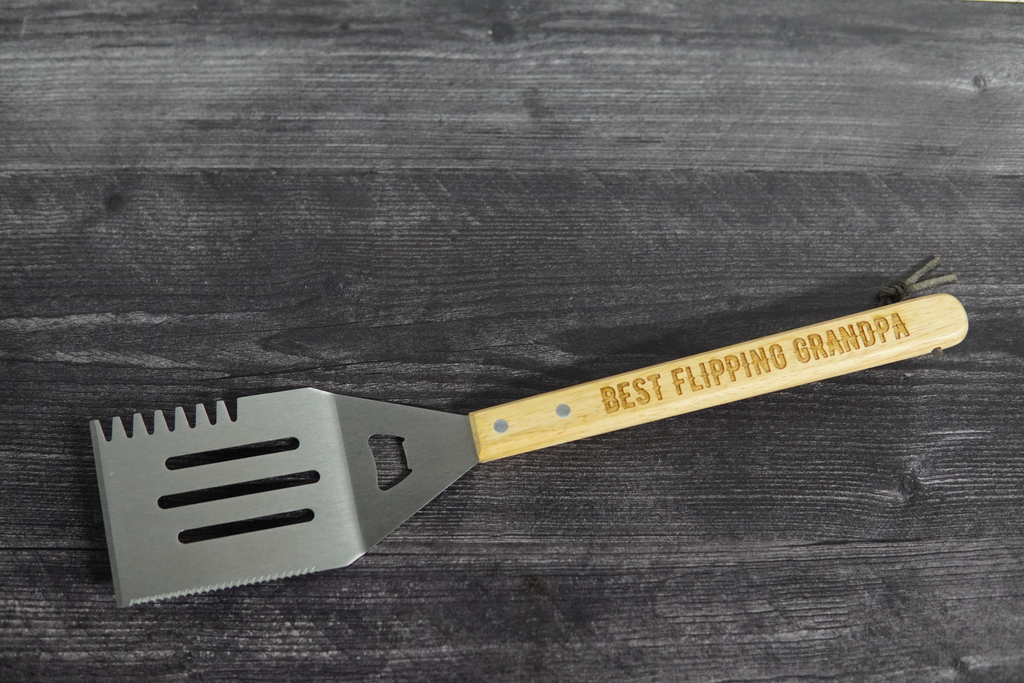 Grill spatula with wooden handle and engraved text on a dark wooden surface