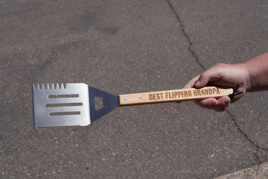 Spatula with 'Best Flipping Grandpa' text held by a hand on a concrete surface